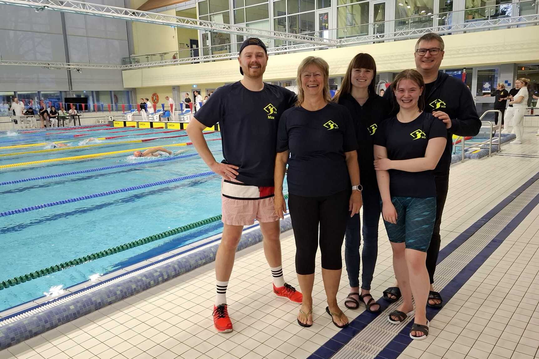 Gruppe von f&uuml;nf Schwimmern in Vereinskleidung steht am Beckenrand eines Schwimmbeckens w&auml;hrend eines Wettkampfs.