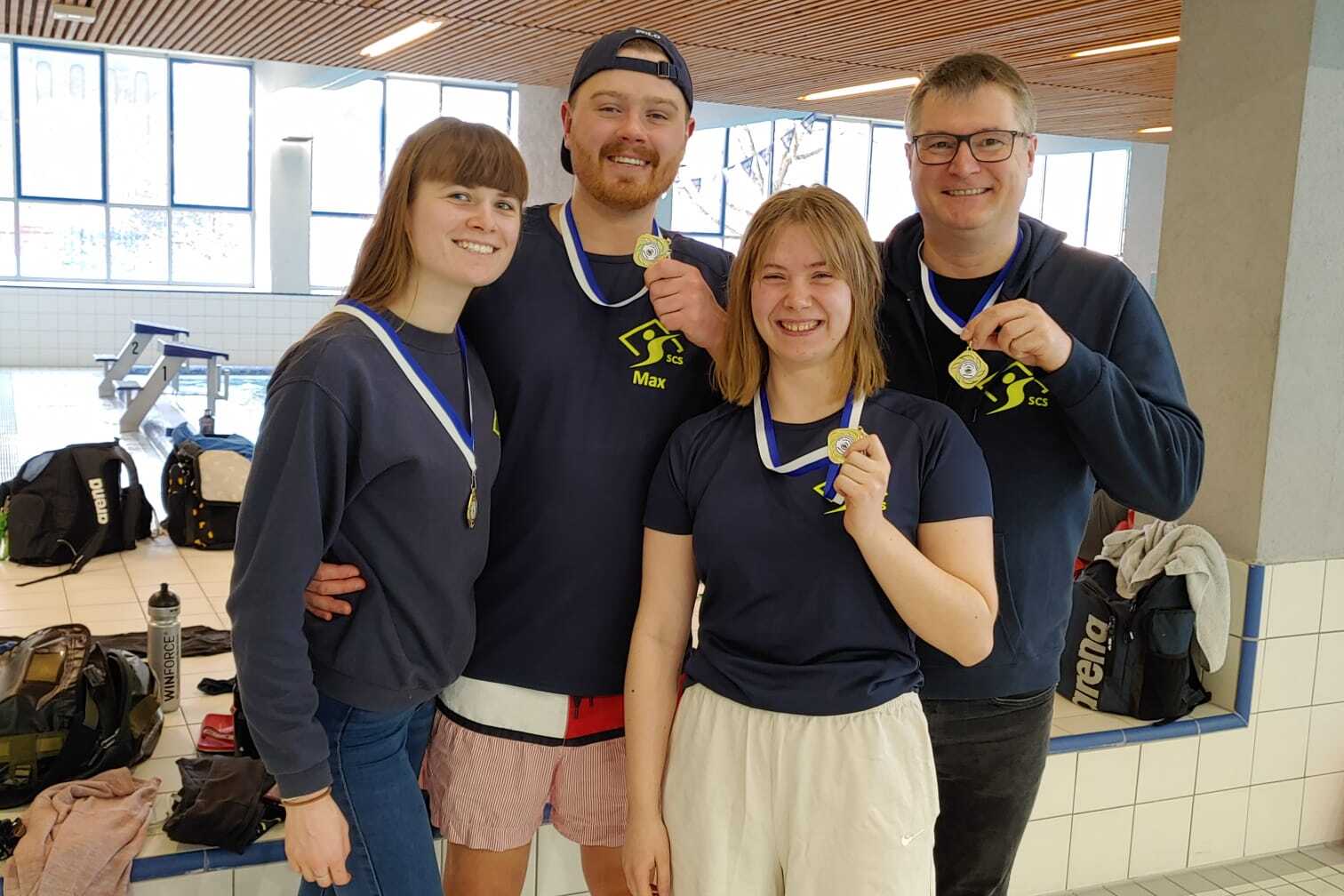 Vier Personen in Sportkleidung halten Medaillen in einer Schwimmhalle, Hintergrund zeigt Schwimmbecken und Taschen.