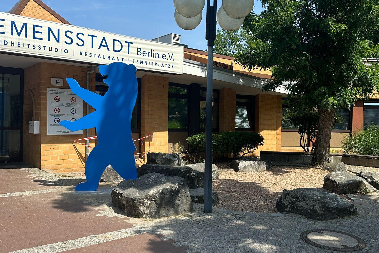 Blaue Figur vor einem Geb&auml;ude mit dem Schriftzug &bdquo;E-SPORTS STADT Berlin e.V.&ldquo; und Laternen im Freien.