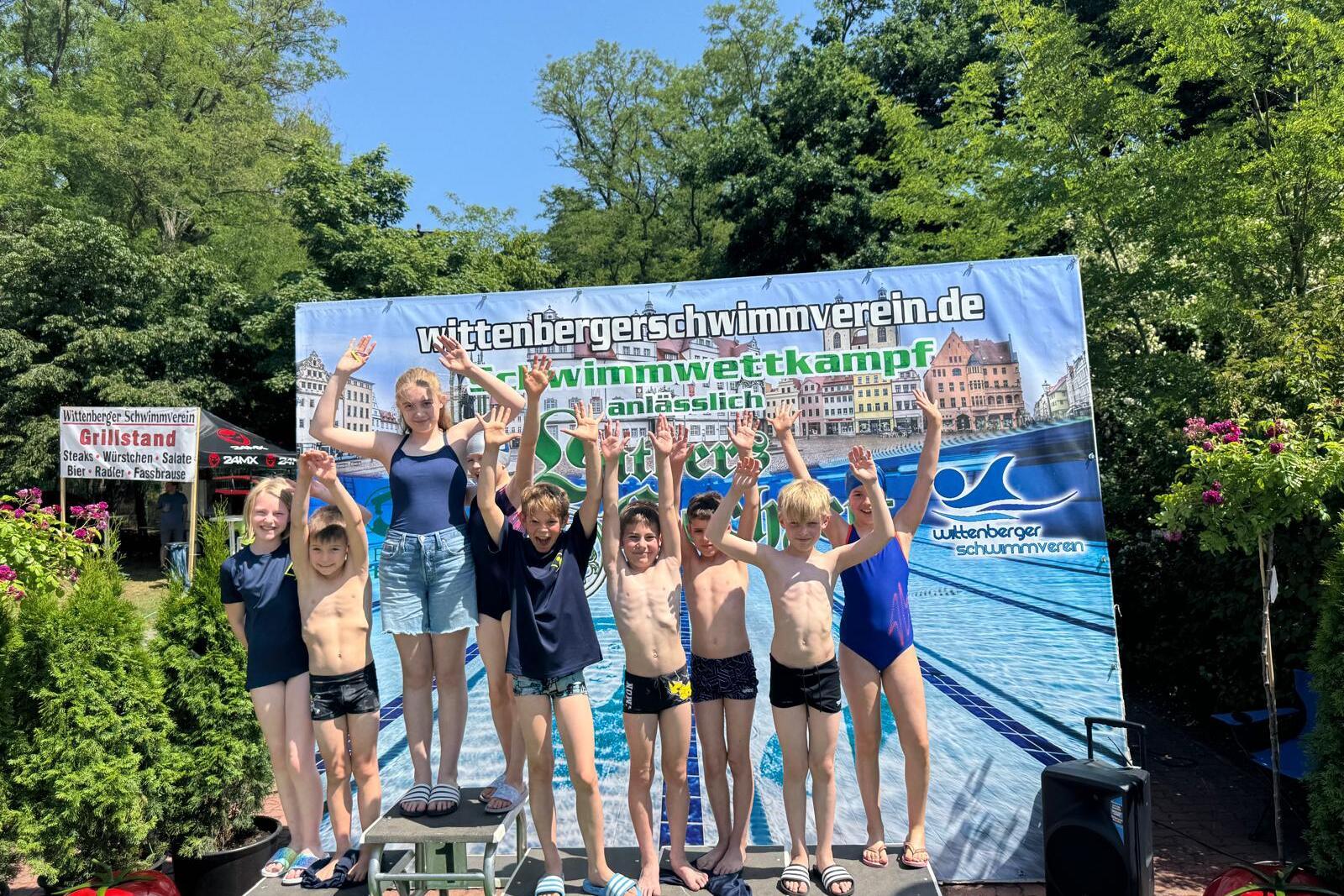 Gruppe von Kindern in Badebekleidung auf einem Siegerpodest, jubelnd vor einem Banner des Wittenberger Schwimmvereins.