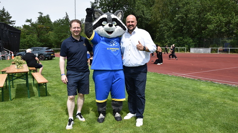 Drei Personen, darunter ein Maskottchen, posieren auf einem Sportplatz mit grüner Wiese im Hintergrund.