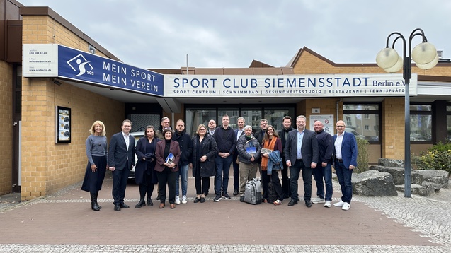 Gruppe von Personen vor dem Eingang des Sport Club Siemensstadt in Berlin, mit Schild und verschiedenen Pflanzen im Hintergrund.