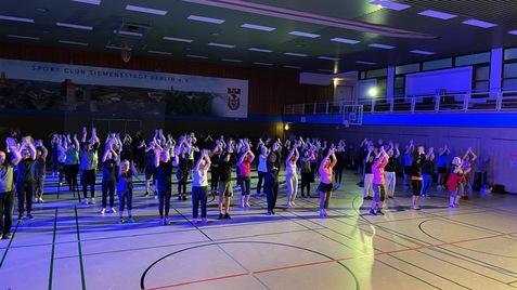Gruppentanzveranstaltung in einer Sporthalle mit Teilnehmern, die synchron ihre Arme in die Luft heben.