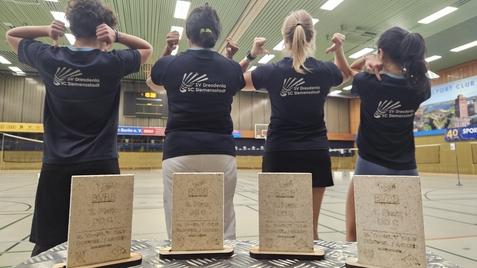 Vier Personen in T-Shirts mit Vereinslogo zeigen ihre Medaillen in einer Sporthalle. Auf einem Tisch stehen drei Pokale.
