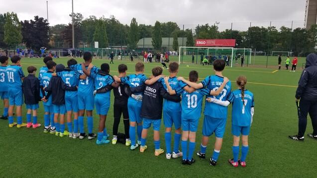 Gruppe von Kindern in blauen Fußballtrikots, die Arm in Arm am Spielfeldrand stehen und ein Spiel beobachten.