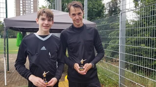 Zwei Fußballspieler in schwarzen Trikots halten Pokale in einer Außenumgebung mit Sportplatz und Zelt im Hintergrund.