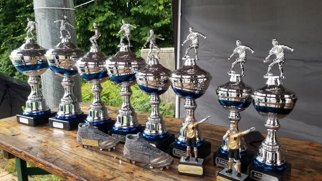 Reihe von silbernen Pokalen mit Figuren und einem Paar Fußballschuhen auf einem Holztisch im Freien.