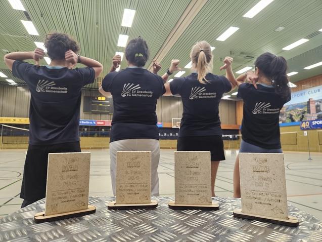 Vier Personen in T-Shirts mit Vereinslogo zeigen ihre Medaillen in einer Sporthalle. Auf einem Tisch stehen drei Pokale.
