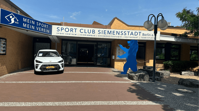 Eingangsbereich des Sport Club Siemensstadt in Berlin mit einem weißen Auto und einer blauen Bärenfigur.