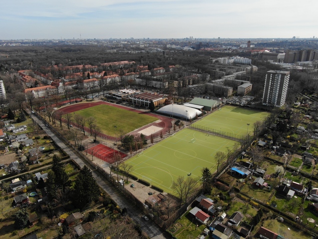 Luftaufnahme eines Sportgeländes mit Tennisplätzen, Fußballfeld und Wohngebäuden in der Umgebung.