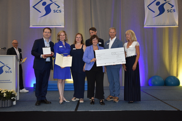 Gruppennahaufnahme von sechs Personen auf einer Bühne, die Auszeichnungen und einen Scheck für ein Projekt präsentieren.