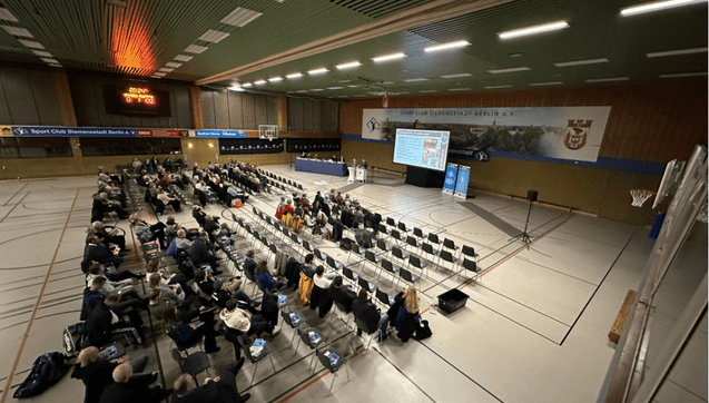 Versammlung in einer Sporthalle mit zahlreichen Stühlen und einer Bühne im Hintergrund. Zuschauer folgen der Präsentation.