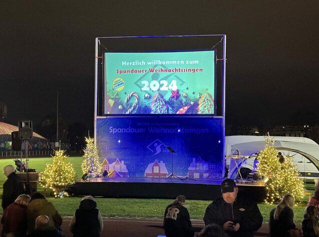 Bühne mit einem digitalen Banner für das Spandauer Weihnachtssingen 2024, umgeben von beleuchteten Tannenbäumen.