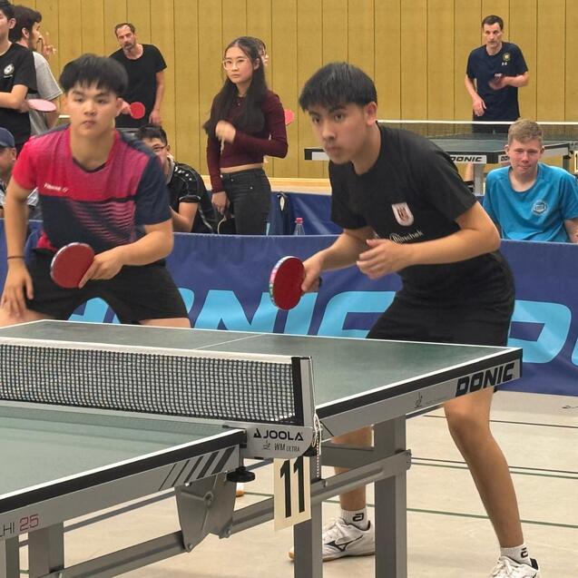 Zwei Tischtennisspieler in Aktion am Tisch, Zuschauer im Hintergrund, Spielatmosph&auml;re bei einem Turnier.