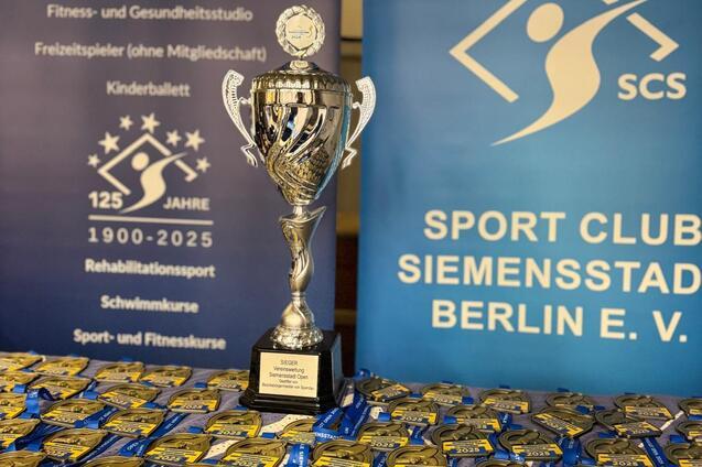 Pokale und Medaillen in einer Sportveranstaltung, im Hintergrund Banner mit Informationen zu Sportangeboten.