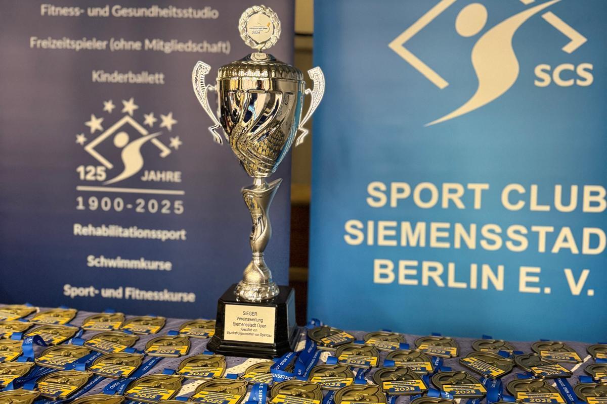 Pokale und Medaillen in einer Sportveranstaltung, im Hintergrund Banner mit Informationen zu Sportangeboten.