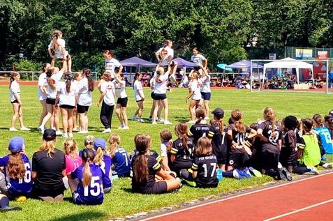 Cheerleadergruppe führt eine Aufführung auf einer Wiese vor zuschauenden Kindern in Sportkleidung durch.