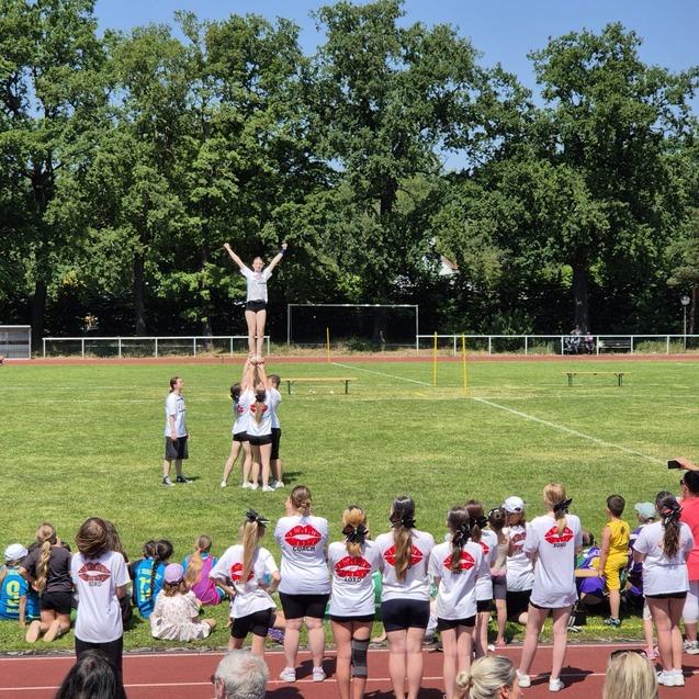 M&auml;dchen in sportlicher Kleidung beim Cheerleading, ein Team hebt eine Person in die H&ouml;he auf einem Sportplatz.