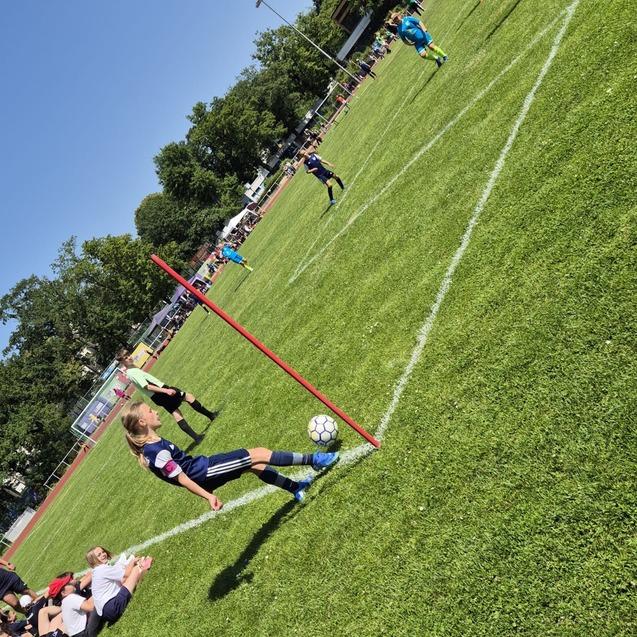 Ein M&auml;dchen f&uuml;hrt einen Eckball auf einem gr&uuml;nen Fu&szlig;ballplatz aus, Zuschauer und Spieler sind im Hintergrund zu sehen.