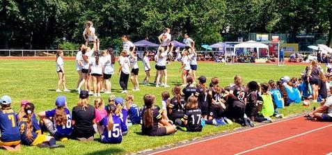 Cheerleadergruppe führt eine Aufführung auf einer Wiese vor zuschauenden Kindern in Sportkleidung durch.