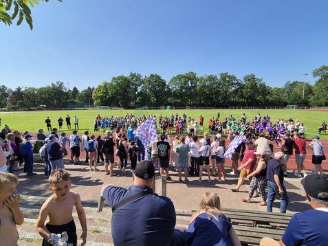 Versammlung von Kindern und Zuschauern auf einem Sportplatz, umgeben von grüner Natur unter blauem Himmel.