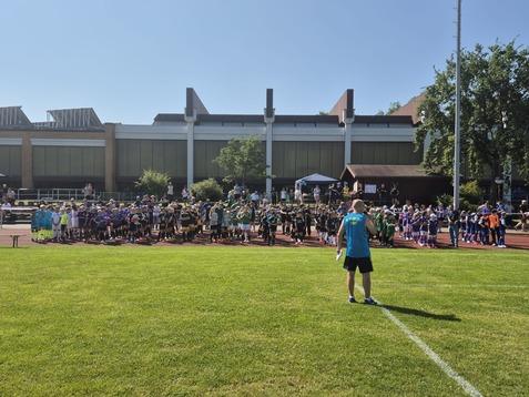 Eine Gruppe von Kindern in Sportkleidung versammelt sich auf einem Rasenplatz, während ein Trainer Anweisungen gibt.