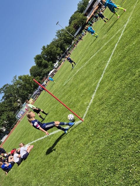 Ein Mädchen führt einen Eckball auf einem grünen Fußballplatz aus, Zuschauer und Spieler sind im Hintergrund zu sehen.