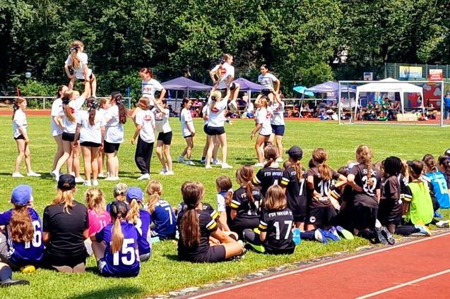Cheerleadergruppe f&uuml;hrt eine Auff&uuml;hrung auf einer Wiese vor zuschauenden Kindern in Sportkleidung durch.