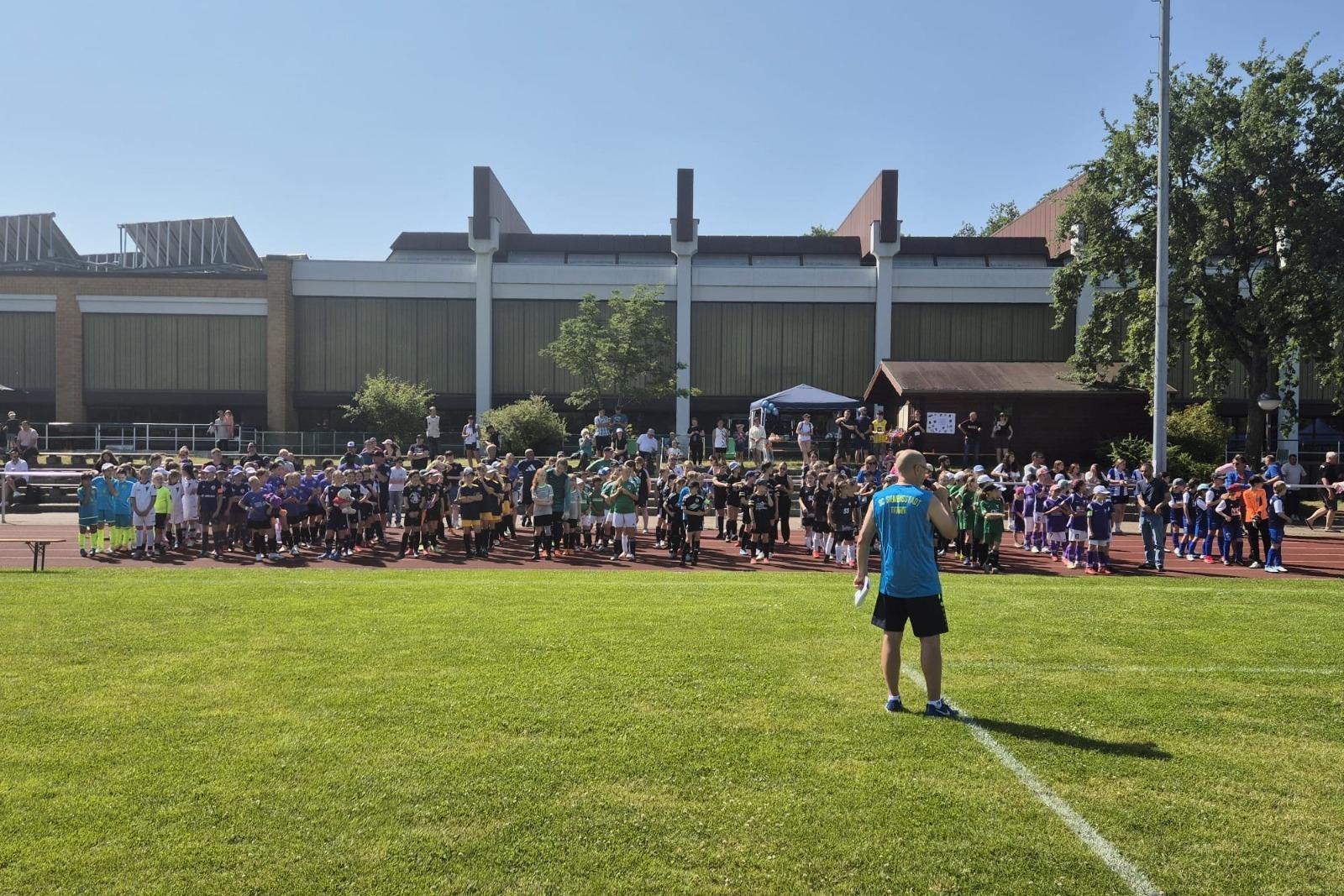Eine Gruppe von Kindern in Sportkleidung versammelt sich auf einem Rasenplatz, w&auml;hrend ein Trainer Anweisungen gibt.