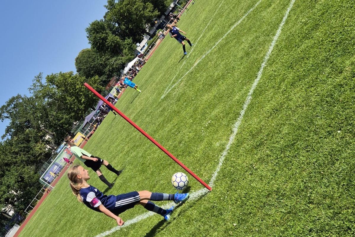 Ein M&auml;dchen f&uuml;hrt einen Eckball auf einem gr&uuml;nen Fu&szlig;ballplatz aus, Zuschauer und Spieler sind im Hintergrund zu sehen.