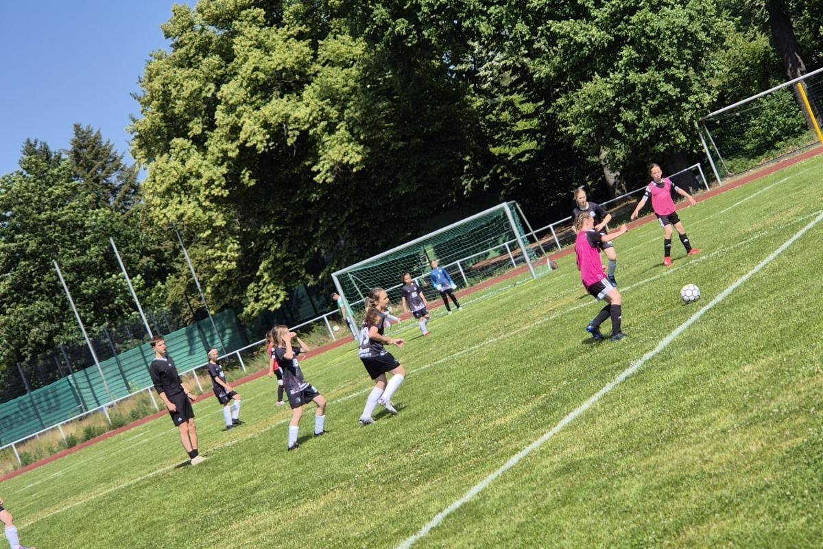 M&auml;dchen beim Fu&szlig;balltraining auf einem gr&uuml;nen Platz, umgeben von B&auml;umen und einem klaren Himmel.