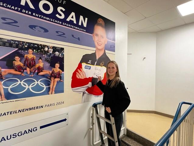 Junge Sportlerin steht neben einem Wandbild mit Olympiateilnehmerinnen der rhythmischen Sportgymnastik.