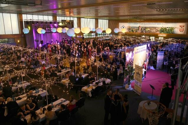 Großes Event mit geschmückten Tischen, tanzenden Gästen und festlicher Beleuchtung in einer Sporthalle.