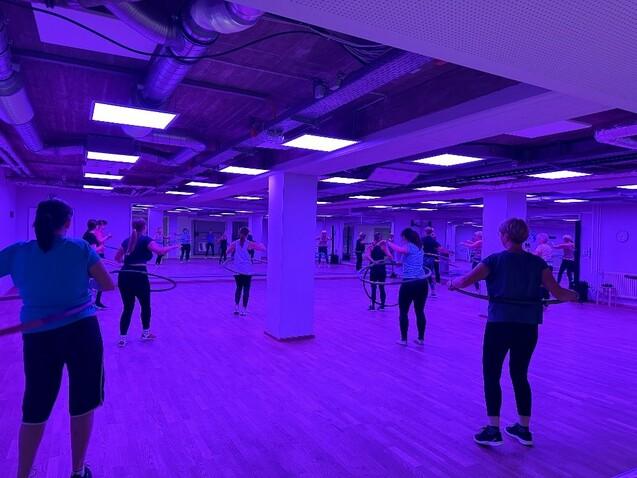 Gruppentraining mit Hula-Hoops in einem beleuchteten Fitnessraum mit violettem Licht und Spiegelwänden.
