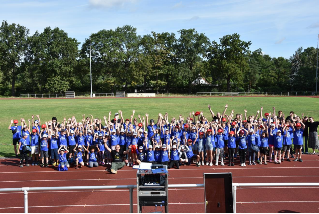 Gruppenfoto von Kindern in blauen T-Shirts, die auf einer Laufbahn stehen und die Hände in die Luft strecken.