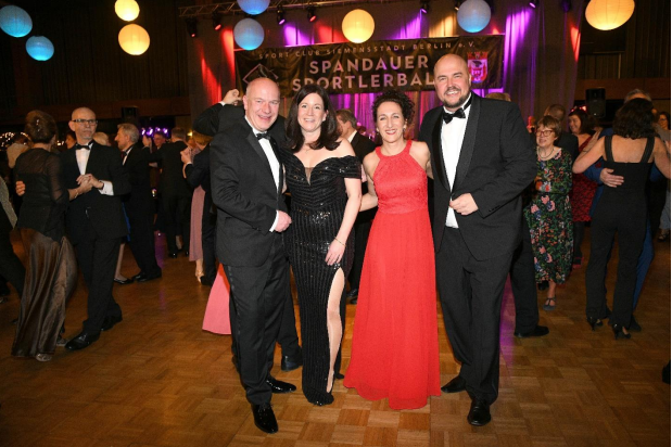 Vier Personen in eleganter Abendgarderobe posieren lächelnd auf einem Ball, mit festlicher Dekoration im Hintergrund.