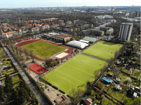 Luftaufnahme eines Sportgeländes mit einem Fußballfeld, einer Leichtathletikbahn und einem angrenzenden Wohngebiet.