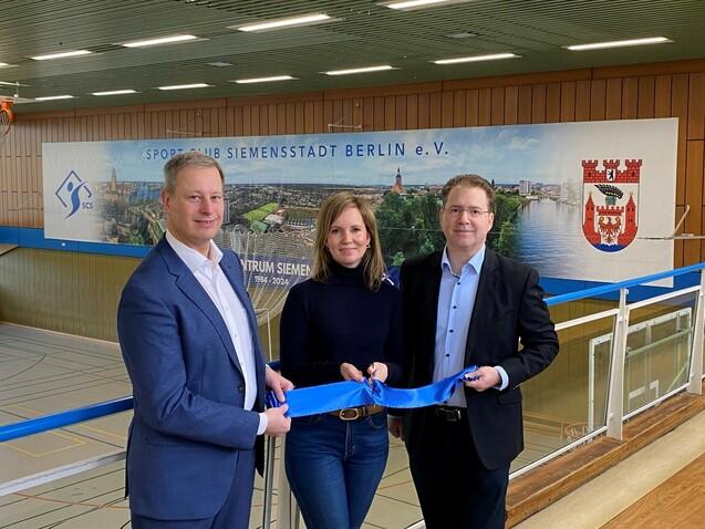 Drei Personen halten ein blaues Band in einer Sporthalle, mit einem Banner des Sport Clubs Siemensstadt im Hintergrund.