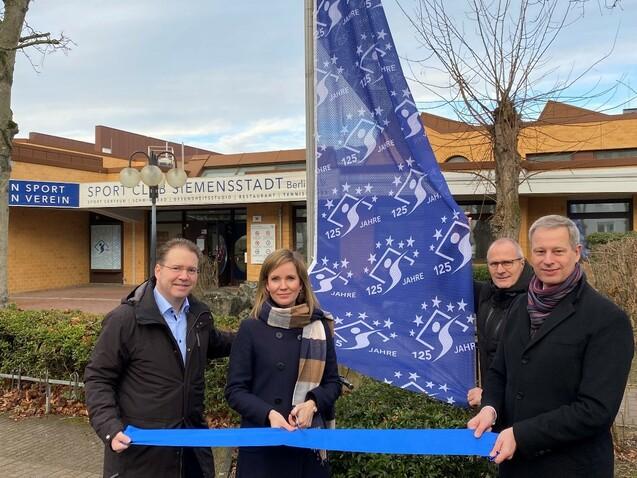 Vier Personen stehen vor dem Sport Club Siemensstadt und halten ein blaues Band, während eine Flagge mit „125 Jahre“ weht.