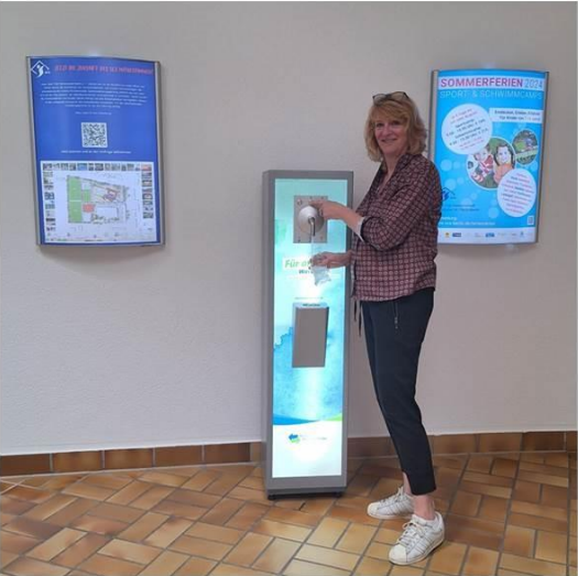 Frau steht an einem Informationsstand und betätigt einen Wasserspender in einem Innenraum.