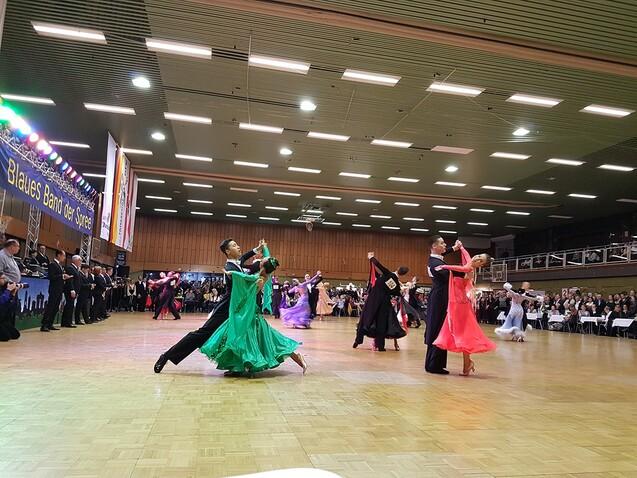 Tanzpaare in eleganter Kleidung tanzen auf einer Bühne bei einem Wettbewerb, Zuschauer im Hintergrund.