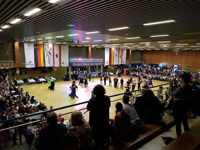 Tanzveranstaltung mit Paaren in bunten Kostümen auf einer Holzfläche, umgeben von Zuschauern und bunten Lichtern.