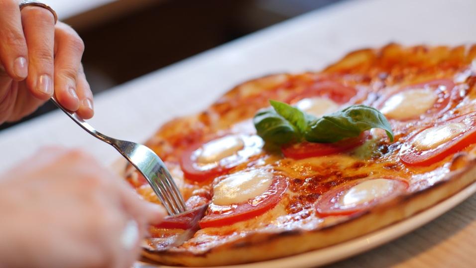 Eine Person schneidet eine Pizza mit Tomaten, Mozzarella und Basilikum auf einem Teller.