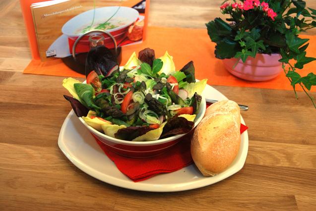 Bunte Salat-Variation mit Gem&uuml;se, Kerne und Br&ouml;tchen auf einem Teller, begleitet von einer Pflanze im Hintergrund.