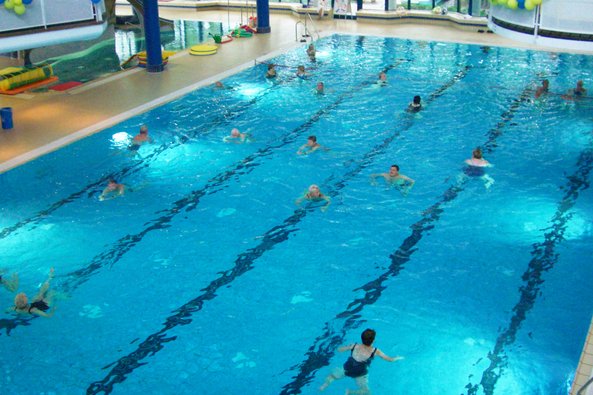 Schwimmbecken mit schwimmenden Personen und blauen und gelben Luftballons an der Decke in einem Innenbereich.