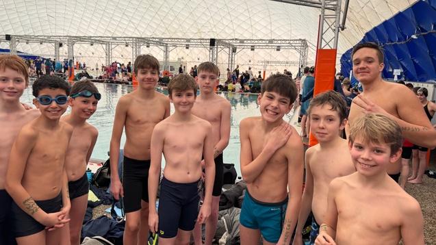 Gruppe von acht Jungen in Badebekleidung, lächelnd, am Rand eines Schwimmbeckens in einer großen Halle.