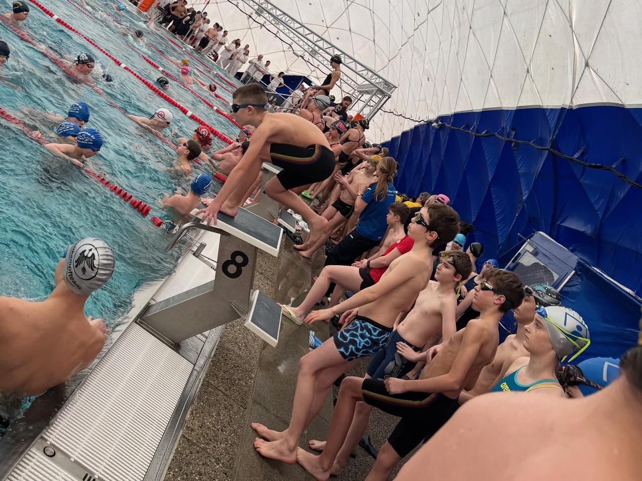 Schwimmer stehen am Beckenrand eines Hallenbads, während andere im Wasser trainieren. Überdachte Umgebung mit Netzen.