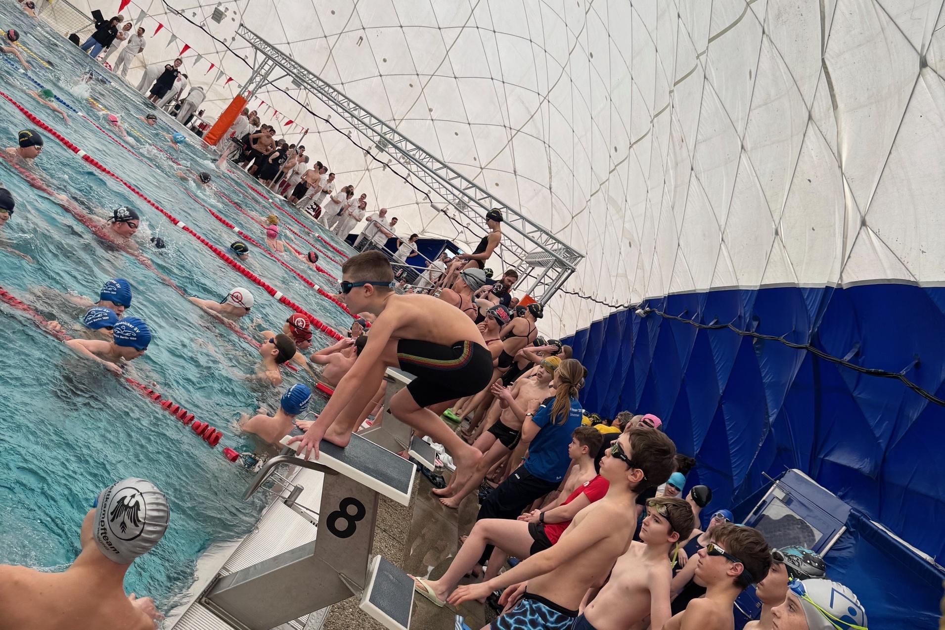 Schwimmer stehen am Beckenrand eines Hallenbads, w&auml;hrend andere im Wasser trainieren. &Uuml;berdachte Umgebung mit Netzen.