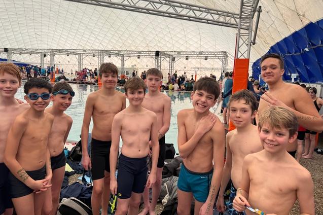 Gruppe von acht Jungen in Badebekleidung, lächelnd, am Rand eines Schwimmbeckens in einer großen Halle.