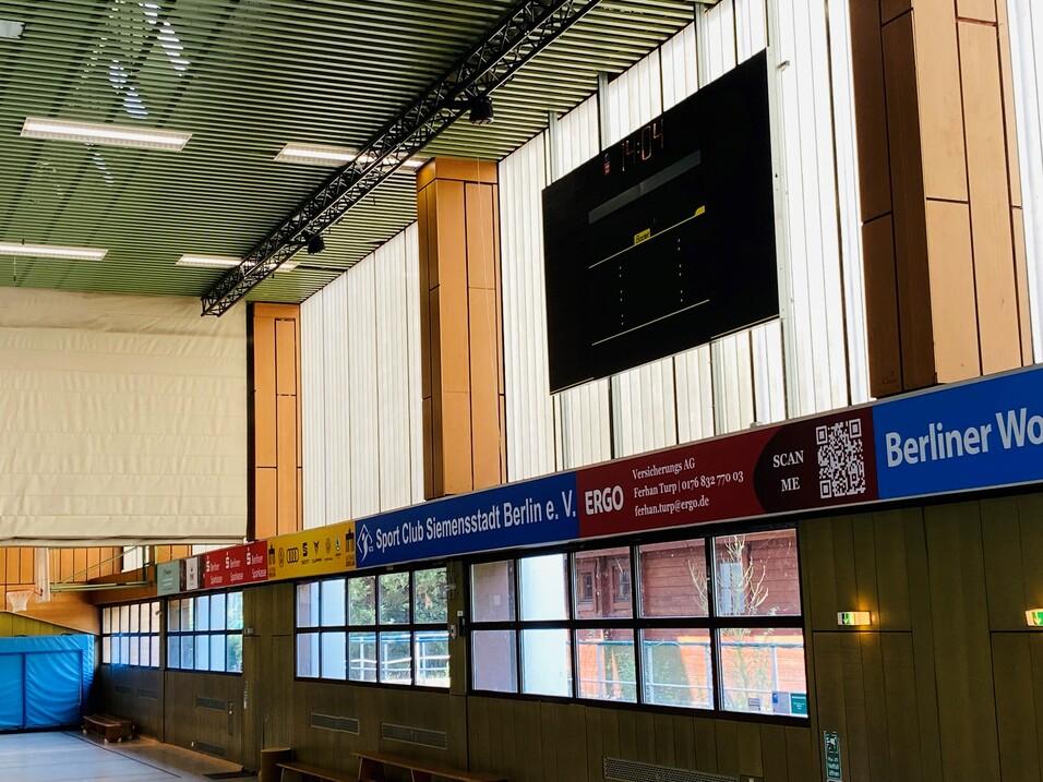 Innenansicht einer Sporthalle mit einem gro&szlig;en Anzeigetafel und bunten Sponsorenbannern an der Wand.